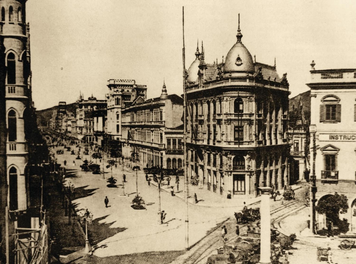 Fotografia histórica em tom sépia do centro do Rio de Janeiro no início do século XX. A imagem mostra uma ampla avenida recém-urbanizada, com calçadas largas e poucas pessoas caminhando ou atravessando a via. À direita, destaca-se um grande edifício de arquitetura eclética, com cúpula e detalhes ornamentais, típico do período. À esquerda, uma sequência de prédios comerciais acompanha o traçado da avenida. A cena transmite a ideia de modernização urbana e transformação da cidade.
