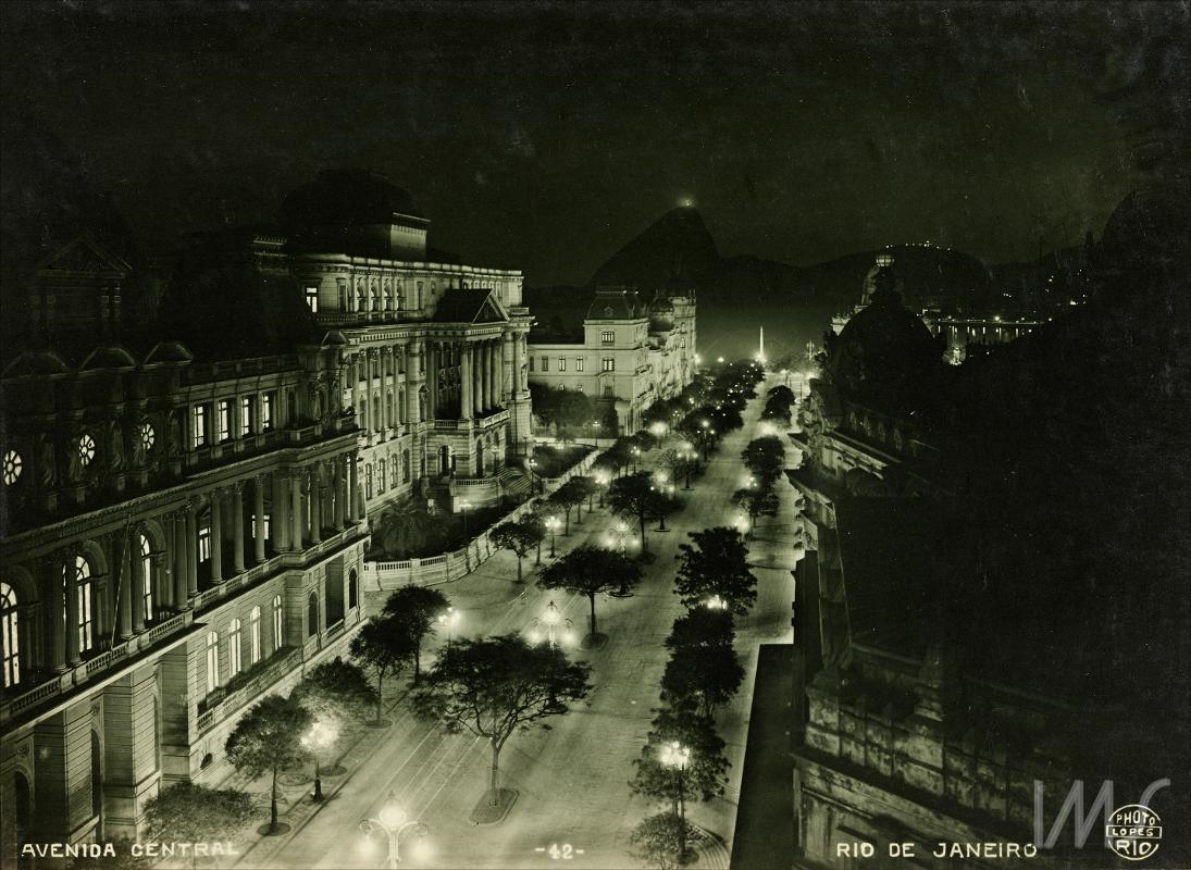 Foto noturna em preto e branco da Av. Central, atual Av. Rio Branco (centro do Rio de Janeiro, início do séc. XX). A imagem, vista de um ponto elevado, mostra a larga avenida iluminada por postes de luz alinhados e árvores no canteiro central. À esquerda e à direita, destacam-se edifícios monumentais de arquitetura eclética, com fachadas ricamente ornamentadas. Ao fundo, surgem morros característicos da paisagem carioca. A cena transmite a modernização urbana e a imponência do Rio naquele período histórico.