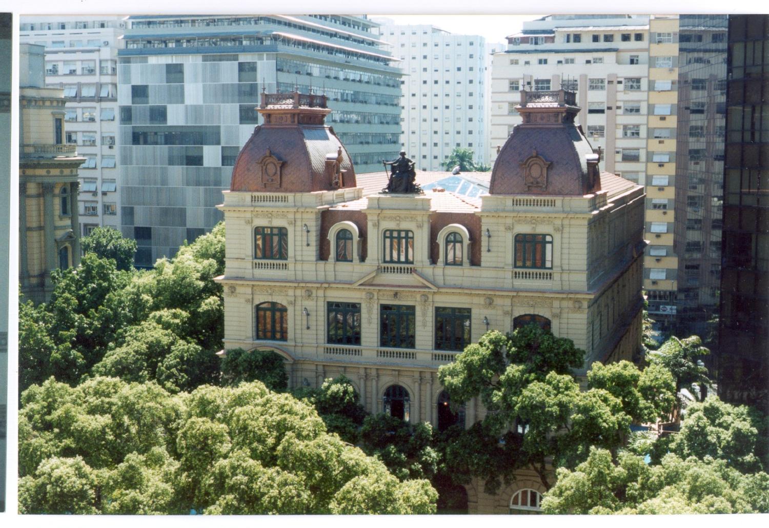 Imagem externa diurna: edifício do CCJF – Centro Cultural Justiça Federal, visto de ponto elevado. Arquitetura eclética, fachada clara e simétrica, com três pavimentos, janelas altas e detalhes ornamentais. No topo, há duas cúpulas laterais em tom avermelhado e, ao centro, uma escultura sobre o telhado. Árvores frondosas cercam a construção em primeiro plano. Ao fundo, aparecem edifícios altos e modernos, criando contraste entre o patrimônio histórico e a paisagem urbana contemporânea do centro do Rio.