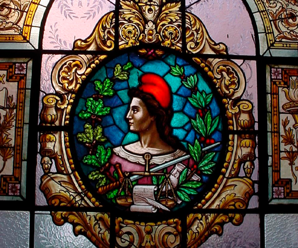 A imagem mostra um vitral colorido com a figura icônica de Marianne, símbolo da República Francesa, usando um gorro frígio vermelho. Ela aparece de perfil, com expressão serena, vestindo uma túnica clara e segurando um ramo verde. Ao redor, há uma moldura circular ricamente decorada com arabescos dourados e elementos florais, em contraste com tons de azul, verde e rosa do fundo. A luz que atravessa o vidro realça os detalhes e confere solenidade à composição.
