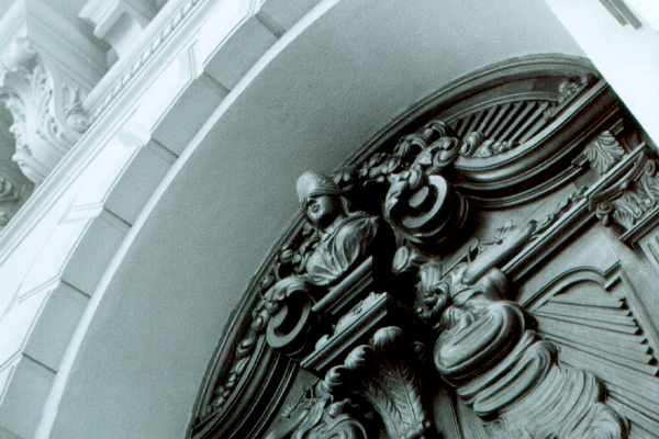 Detalhe em preto e branco da porta principal do CCJF – Centro Cultural Justiça Federal. A imagem mostra uma grande porta de madeira ricamente entalhada, com ornamentos clássicos e uma figura decorativa no centro, integrada ao desenho da madeira. A porta é emoldurada por um arco de pedra com elementos arquitetônicos elaborados, como relevos e molduras curvas. O enquadramento em ângulo enfatiza a altura e a imponência da entrada, destacando o caráter histórico e institucional do edifício.