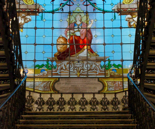 Escadaria principal do CCJF – Centro Cultural Justiça Federal. Em 1º plano, vê-se a escada com degraus de pedra e corrimãos laterais em metal trabalhado, criando um enquadramento simétrico. Ao fundo, há um grande vitral colorido que representa a Deusa da Justiça, sentada, com vestes em tons de vermelho e dourado, segurando símbolos associados à justiça. O vitral é composto por desenhos ornamentais e molduras decorativas, permitindo a entrada de luz natural e reforçando o caráter histórico e solene do espaço
