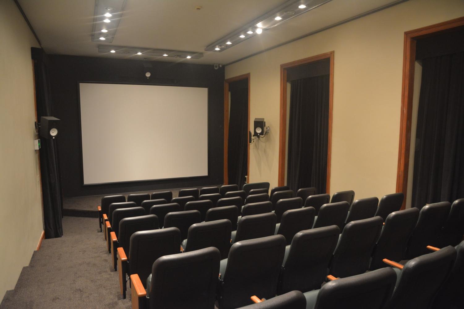 A foto mostra um auditório pela visão do público, com um telão na frente. Há cadeiras pretas com detalhes em madeira, uma mesa central em cima do tablado, também com cadeiras. As paredes são beges e há cortinas pretas nas janelas. O ambiente é bem iluminado. É o cinema do Centro Cultural Justiça Federal.