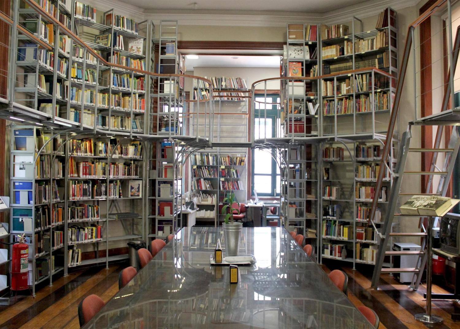 Imagem da biblioteca do CCJF – Centro Cultural Justiça Federal. O espaço é amplo e organizado, com estantes altas de madeira repletas de livros dos dois lados, indo do chão ao teto. Escadas metálicas móveis permitem acesso às prateleiras superiores. No centro da sala, há uma longa mesa retangular com superfície brilhante, cercada por cadeiras, usada para leitura e estudo. O piso é de madeira e, ao fundo, janelas altas permitem a entrada de luz natural, criando um ambiente silencioso e acolhedor.