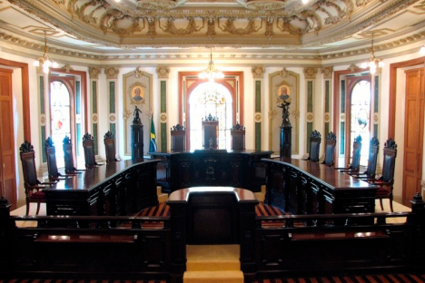 A foto ilustra a Sala de Sessões do Centro Cultural Justiça Federal, localizado na Cinelândia, na cidade do Rio de Janeiro. Nela, há móveis no estilo colonial, mesa em formato de ‘U’, e cadeiras altas entalhadas, formando um parlamento do judiciário. O local foi a sede do Supremo Tribunal Federal até 1960.