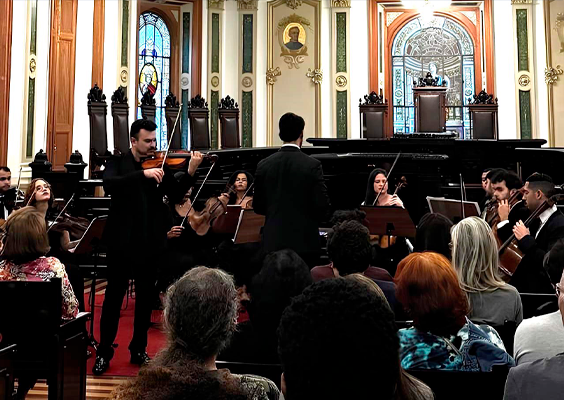 Na foto, um grupo de musicistas, entre homens e mulheres, tocam violino e outros instrumentos de corda. Eles se apresentam para um público diverso, que aparece de costas. No plano de fundo, mobiliários como bancadas e cadeiras, no estilo colonial, de madeira escura. As paredes e janelas tem pinturas de época, com detalhes em arabescos.