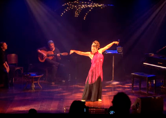 A foto mostra uma mulher branca de cabelos presos, enrolados, dançando em um palco. Ela está com os braços abertos, esticados para os lados. Usa vestido longo preto e uma blusa com franjas, rosa, por cima. Ao fundo, dois músicos, um tocando violão e o outro instrumento de percussão. Do lado direito, há parte de um piano de cauda preto. Acima, no teto, pequenas luzes pisca-piscas e a iluminação do palco em tons de azul e amarelo.