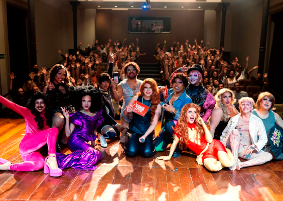 Fotografia de um grupo de drags queens, com vestimentas coloridas e brilhantes, sentadas em um palco de madeira. Atrás dela, uma plateia cheia com as mãos levantadas para cima. Todos parecem animados. 