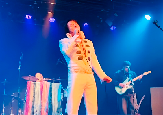 A foto mostra um homem branco de cabelos pretos, que segura um microfone na mão e canta. Ele está inclinado levemente para trás. Ele usa blusa e calça brancas, com detalhes dourados, assemelhando aos trajes usados pelo Rei do Rock, Elvis Presley. Atrás dele, dois outros músicos, um tocando violão e outro, bateria. A iluminação do palco é azul.