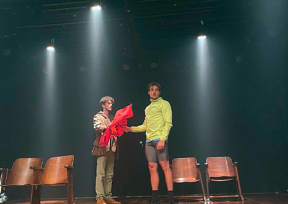 Imagem de dois homens brancos de cabelos castanhos contracenando, em um palco. Um deles segura um guarda-chuva vermelho nas mãos. Ele veste casaco de manga comprida verde claro e bermuda ciclista cinza. O outro usa casaco marrom e calça cáqui. Nas laterais do cenário, quatro cadeiras de madeira, duas de cada lado. Há três spots de luz iluminando o local.