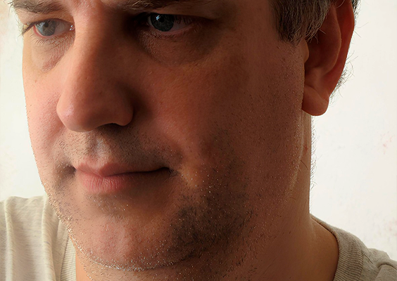 Fotografia. Close-up no rosto de um homem de cabelo grisalho e olhos claros. O homem está com a barba por fazer e usa camisa de malha bege clara. Ele está sério e pensativo e olhando ligeiramente para o lado.