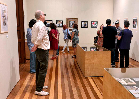 Exposição em galeria. Pessoas circulam salão com paredes bege e quadros emoldurados em diversos padrões diferentes, pendurados. Ao centro, há dois expositores em madeira clara e tampo de vidro pelo qual avistamos álbuns abertos. Senhor, ligeiramente calvo e de cabelos grisalhos, de óculos, olha para os visitantes da exposição. Estes estão posicionados ao fundo da sala: homens e mulheres de meia idade. A maioria olha os quadros e há um grupo que conversa enquanto uma mulher os fotografa.