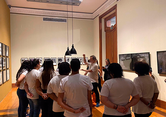 A foto mostra um grupo de pessoas, a maioria mulheres, de costas. Elas usam blusa branca e calça jeans. No centro, mais ao fundo, um homem branco com camisa clara e calça escura tem um dos braços para o alto e o outro na altura do peito, como se tivesse demonstrando alguma explicação. Nas paredes beges, quadros com obras de arte. Ao fundo, do lado direito, uma grande porta de madeira.