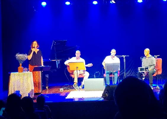 A imagem mostra um grupo de quatro pessoas em um palco iluminado por luzes azuis. Uma mulher à esquerda está em pé, segurando um microfone, enquanto três homens estão sentados tocando instrumentos: uma guitarra, uma flauta e uma percussão. Há um piano de cauda ao fundo, embora não esteja sendo tocado. À esquerda, sobre uma mesa coberta com uma toalha, há um arranjo decorativo. A plateia é parcialmente visível nas sombras, sugerindo que a imagem foi tirada durante uma apresentação musical ao vivo.