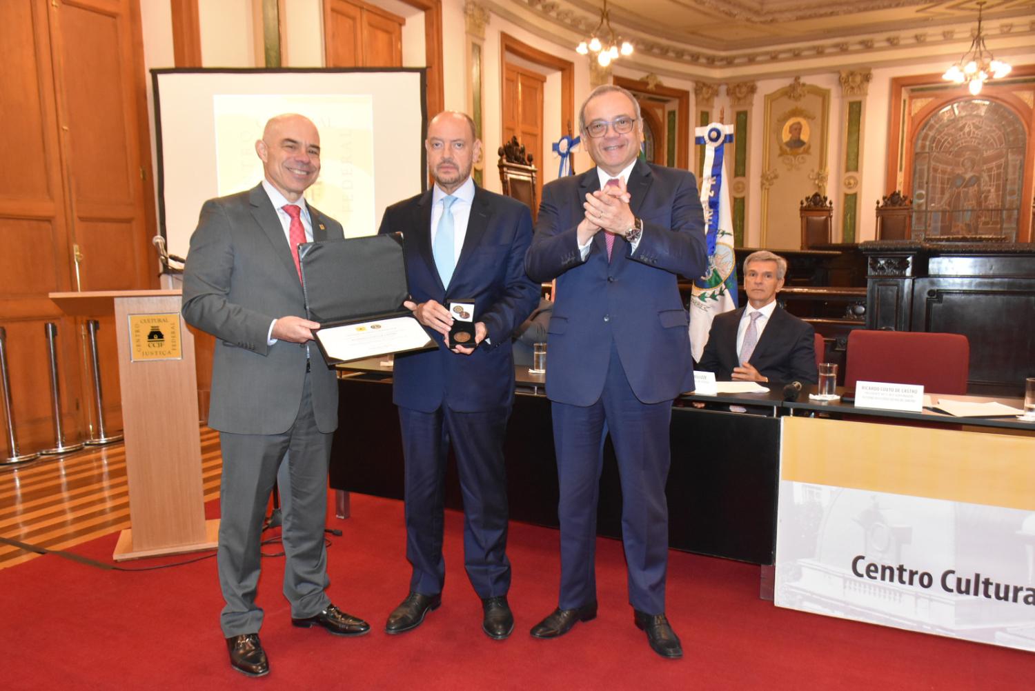 Fotografia realizada na Sala de Sessões do Centro Cultural Justiça Federal. Em primeiro plano, três homens de terno participam de uma cerimônia de homenagem. À esquerda, um homem sorri enquanto exibe um diploma em estojo aberto. Ao centro, outro segura uma medalha em caixa de apresentação. À direita, um terceiro homem aplaude. Ao fundo, vê-se a mesa de autoridades, bandeiras, vitrais históricos e a arquitetura ornamentada do salão. O ambiente é solene e celebra uma distinção institucional.
