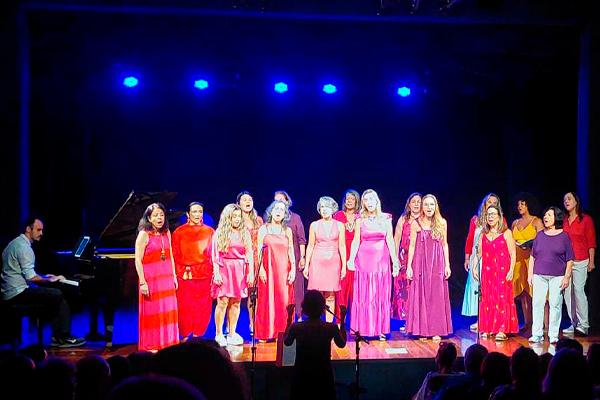 Grupo de mulheres se apresenta no palco de um teatro, sob luzes azuis e iluminação frontal. A maioria usa roupas em tons de rosa e vermelho. Uma pessoa conduz o grupo da plateia, que está cheia e em silêncio. A cena é vista do fundo do auditório, com foco na performance musical.