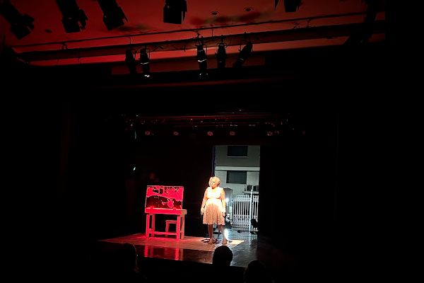 Cena teatral com iluminação focada em uma atriz no palco, vestida com roupa clara. Ela está em pé ao lado de um móvel vermelho com detalhes em preto, enquanto todo o restante do ambiente permanece escuro. A luz destaca apenas a atriz e parte do cenário, criando um clima dramático e intimista.