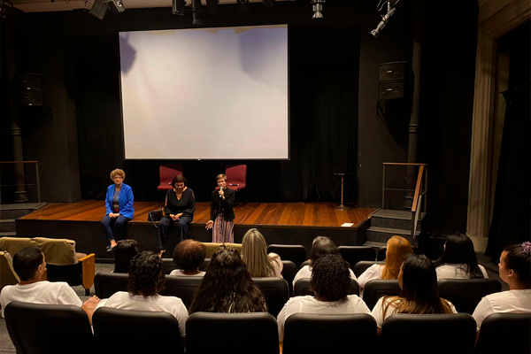 A foto mostra um auditório com uma tela de cinema e um pequeno palco. Nele, três mulheres brancas estão sentadas, uma delas diz algo, com o microfone na mão. Na plateia, mulheres de blusa branca estão sentadas, assistindo. As poltronas são pretas.