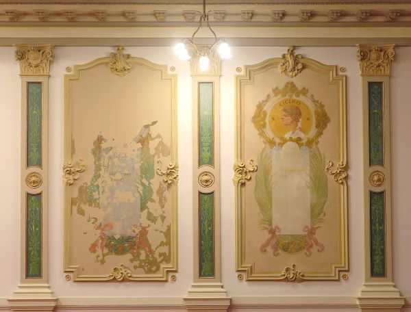 Parede interna ornamentada da Sala de Sessões do Centro Cultural Justiça Federal, com duas grandes pinturas emolduradas lado a lado. À esquerda, a pintura está bastante desgastada, com grandes áreas descascadas. À direita, a imagem está preservada e mostra o busto do orador romano Cícero acima de uma inscrição apagada, ladeada por folhas de louro e enfeites em rosa e dourado. Ao redor, detalhes decorativos em dourado e verde.