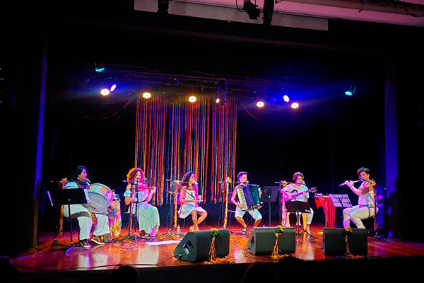 Apresentação musical no palco com sete mulheres tocando instrumentos como zabumba, triângulo, sanfona, flauta e violão. Todas estão vestidas com roupas claras, e o fundo é decorado com fitas coloridas penduradas. A iluminação cênica mistura tons quentes e frios, criando um clima festivo e acolhedor típico de festas populares.