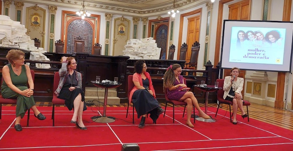 Cena teatral com iluminação focada em uma atriz no palco, vestida com roupa clara. Ela está em pé ao lado de um móvel vermelho com detalhes em preto, enquanto todo o restante do ambiente permanece escuro. A luz destaca apenas a atriz e parte do cenário, criando um clima dramático e intimista.