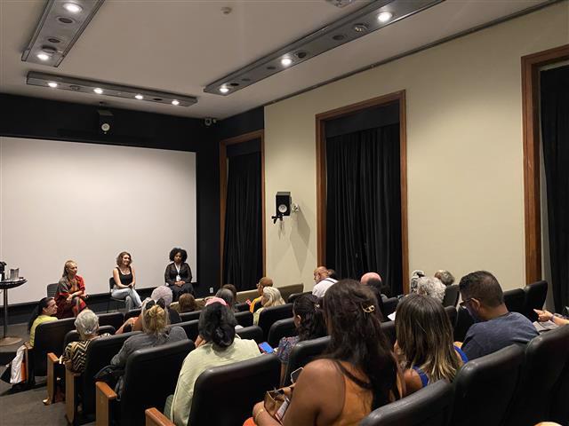 Na imagem, o cinema do CCJF com poltronas ocupadas por um público atento. Na frente, três mulheres estão sentadas em cadeiras, participando de uma conversa ou roda de debate diante de uma tela branca de projeção. O ambiente é acolhedor, com iluminação no teto e cortinas escuras nas janelas laterais.
