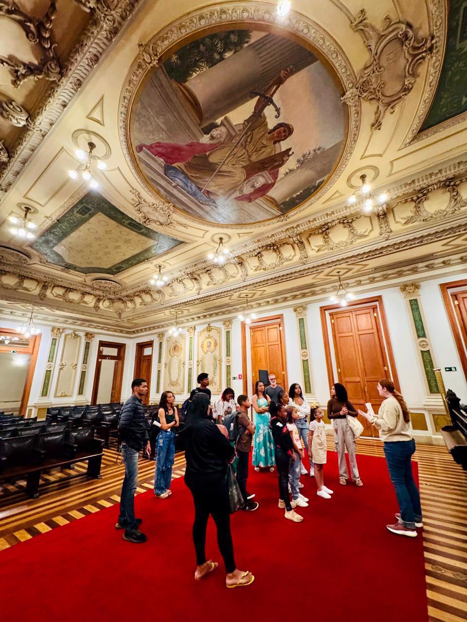 Um grupo de pessoas, incluindo crianças e adultos, está reunido sobre um tapete vermelho em uma sala histórica de teto ornamentado e afrescos. A guia, de blusa clara, fala ao grupo, que a escuta atentamente. O espaço tem portas de madeira altas, paredes decoradas e poltronas pretas dispostas em fileiras, sugerindo um ambiente de visita cultural em um prédio antigo e elegante.