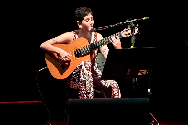 Pessoa sentada em um palco escuro, tocando violão e cantando ao microfone. Ela usa uma roupa estampada e está concentrada na apresentação. Há partituras à sua frente e equipamentos de som ao redor. A iluminação foca apenas nela, destacando a performance musical em um ambiente intimista.