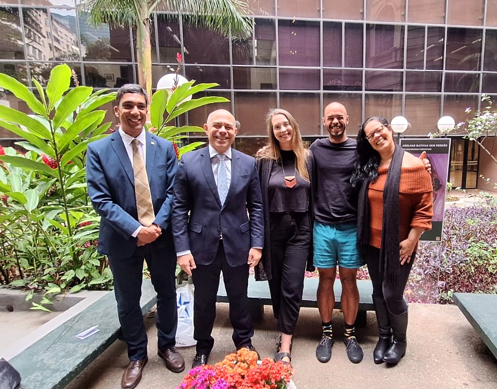 Cinco pessoas posam sorridentes em um jardim, em frente a um prédio envidraçado. À esquerda, dois homens usam terno e gravata. Ao centro, uma mulher veste roupa preta e colar geométrico, ao lado de um homem de camiseta preta e bermuda azul. À direita, uma mulher usa blusa marrom de ombro caído, cachecol preto e botas. À frente, há flores coloridas.