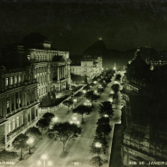 Foto noturna em preto e branco da Av. Central, atual Av. Rio Branco (centro do Rio de Janeiro, início do séc. XX). A imagem, vista de um ponto elevado, mostra a larga avenida iluminada por postes de luz alinhados e árvores no canteiro central. À esquerda e à direita, destacam-se edifícios monumentais de arquitetura eclética, com fachadas ricamente ornamentadas. Ao fundo, surgem morros característicos da paisagem carioca. A cena transmite a modernização urbana e a imponência do Rio naquele período histórico.
