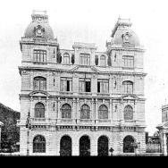 Fotografia histórica em preto e branco do edifício do Supremo Tribunal Federal (STF) quando ainda funcionava no Rio de Janeiro, antes da transferência para Brasília. A imagem mostra um prédio imponente de arquitetura eclética, com três pavimentos, fachada simétrica, janelas em arco e rica ornamentação. Duas torres laterais com relógios coroam a construção. Em primeiro plano, vê-se a área externa do edifício, com construções menores ao redor. A imagem transmite solenidade e importância institucional.