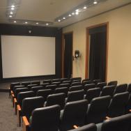 A foto mostra um auditório pela visão do público, com um telão na frente. Há cadeiras pretas com detalhes em madeira, uma mesa central em cima do tablado, também com cadeiras. As paredes são beges e há cortinas pretas nas janelas. O ambiente é bem iluminado. É o cinema do Centro Cultural Justiça Federal.
