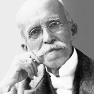 A imagem apresenta um retrato em preto e branco de Ruy Barbosa, já idoso, com expressão séria e contemplativa. Ele usa óculos de aro fino, tem bigode espesso e cabelos ralos e claros. Está vestido com traje formal, incluindo paletó e camisa de colarinho alto, apoiando o rosto levemente sobre uma das mãos, o que reforça a impressão de intelectualidade e reflexão.