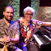 Duas pessoas sorrindo, sentadas ao lado de um piano. Da esquerda para a direita: homem com barba e cabelos curtos escuros segura instrumento de percussão. Ele usa camisa de manga curta com estampas geométricas; homem mais velho, de cabelos brancos, óculos e camisa estampada com cores vibrantes. Ele está sentado ao piano, com a mão esquerda repousando sobre o instrumento. Ao fundo, uma parede de pedra com iluminação suave, sugerindo que estão em um local de apresentação musical, como um palco ou estúdio.