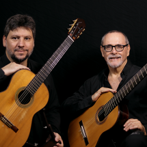 A imagem mostra dois homens sentados, cada um segurando um violão clássico. Ambos vestem camisas pretas e estão de frente para a câmera, com expressões serenas. O homem à esquerda tem barba curta e cabelo castanho, enquanto o da direita usa óculos, tem barba grisalha e está sorrindo. O fundo é preto, destacando os músicos e seus violões. A imagem transmite um clima de seriedade e paixão pela música.