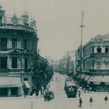 Fotografia antiga do Largo da Carioca, Rio de Janeiro. Os edifícios em destaque possuem arquitetura elaborada com ornamentos detalhados nas fachadas, característica do estilo europeu do início do século XX. A rua é movimentada, com pessoas caminhando e alguns veículos de tração animal. Há também uma linha de bonde no centro da via. O cenário transmite uma atmosfera clássica e urbana, com foco na vida cotidiana da época.