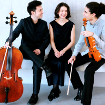 Na foto, dois homens e uma mulher branca estão sentados em um banco; todos sorriem. Um deles (à esquerda) usa paletó preto e segura um violoncelo. A mulher, no meio dos dois, veste vestido longo preto e olha para um dos musicistas. O homem da direita usa blusa social clara e calça preta; ele segura um violino.