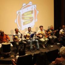 Um grupo de músicos, se apresentam em um palco. Todos estão sentados, tocando e cantando. Entre os instrumentos musicais, estão: pandeiro, banjo, violão, cavaquinho e tan tan.