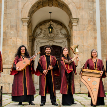 A imagem mostra um grupo de seis músicos, todos vestidos com trajes tradicionais, longos e em tons de vinho com detalhes dourados. Eles estão posando em frente a um edifício antigo, que parece ser uma igreja com uma arquitetura clássica de pedra. Cada um está segurando um instrumento musical antigo: alaúdes, flauta, um órgão portátil, uma harpa e um par de sinos. O grupo aparenta ser especializado em música antiga ou medieval, e o cenário reforça essa atmosfera histórica e cultural.