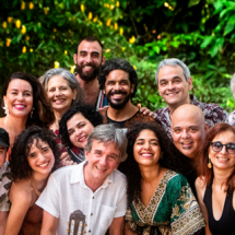 Na foto, um grupo de 15 pessoas, entre homens e mulheres das mais variadas faixas etárias, sorri para a câmera. Eles estão em uma área aberta, com vegetação ao fundo. Há mulheres de cabelos castanhos lisos e enrolados, ruiva, e grisalhas; homens de cabelos castanhos curtos, castanhos enrolados, careca e grisalhos. As roupas se misturam entre estampadas e lisas (brancas e pretas). É possível perceber a ponta de um violão na mão de um dos homens que está posicionado ao centro, na fileira da frente da foto.