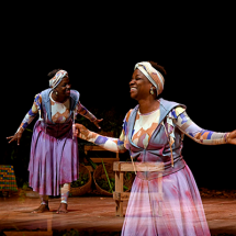 A imagem mostra um palco com uma mulher negra sorridente, descalça, usando vestido longos roxos e faixa de cabeça combinando – parece estar em uma performance teatral, com gestos abertos e expressões alegres. Esta mesma imagem da mulher é espelhada e sobreposta em um primeiro plano da fotografia, com certa transparência. O cenário tem um fundo escuro e alguns objetos de madeira, sugerindo uma ambientação simples e natural. Há uma sensação de movimento e energia, destacando a leveza e o dinamismo da cena.