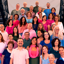 A imagem mostra um grande grupo de pessoas posando na escadaria do CCJF, formando várias fileiras. Eles estão vestidos com roupas coloridas, predominantemente em tons de rosa, azul, verde e roxo, e todos parecem sorrir para a câmera, transmitindo uma atmosfera alegre e amistosa. As idades dos participantes variam, incluindo adultos de meia-idade e idosos.