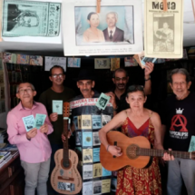 Grupo com 5 homens e uma mulher brancos, em sala com livretos de literatura de cordel. Um deles veste traje plástico transparente com livretos amarelos, azuis e brancos, de cordel. Usa chapéu preto e tem um violão em uma das mãos. Uma mulher de óculos, ao centro, toca outro violão e veste vestido estampado na cor vinho. Os outros homens seguram livretos de cordel, alguns usam óculos e alguns possuem cabelos grisalhos curtos. No 1º plano, na parte superior da imagem, há 5 livros do tema pendurados em varal.