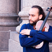 A imagem mostra um homem com cabelo preso e barba, usando uma camisa azul, abraçando um violino. Ele olha para o lado, com uma expressão contemplativa. Ao fundo, há uma estrutura de pedra, sugerindo que ele está em um local histórico ou cultural. A cena transmite uma sensação de apreço pela música e pelo instrumento, em um ambiente tranquilo e introspectivo.