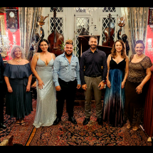 Grupo de pessoas posando em um ambiente elegante e decorado de forma clássica, com paredes vermelhas, lustres, quadros e mobília detalhada. Todos estão vestidos formalmente, com trajes de gala como vestidos longos e ternos, sugerindo que estão participando de um evento especial ou cerimônia. O ambiente tem um tapete com padrão floral e móveis sofisticados, criando uma atmosfera luxuosa e histórica. Ao fundo, há detalhes arquitetônicos, como janelas em estilo gótico e estatuetas decorativas