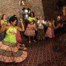 A imagem mostra um grupo de pessoas participando de uma dança tradicional em um ambiente rústico. A maioria das mulheres veste saias coloridas e estampadas, e algumas usam camisetas verdes com o nome "Zanzar", sugerindo uma possível organização cultural. Elas estão dançando e sorrindo, criando um ambiente de alegria e descontração. O piso de azulejos e as paredes de pedra acrescentam um toque autêntico ao local, evocando uma atmosfera de celebração da cultura popular.