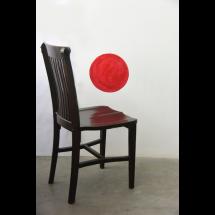Uma cadeira de madeira escura com encosto alto está posicionada contra uma parede branca. Sobre a parede, um círculo vermelho pintado parece flutuar ao lado da cadeira. A superfície do assento reflete levemente o tom do círculo, criando um efeito visual intrigante. O chão é simples e neutro, sem outros elementos ao redor. A composição minimalista sugere um conceito artístico ou simbólico.