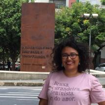 A imagem mostra uma mulher sorridente de cabelos cacheados e óculos, vestindo uma camiseta rosa com os dizeres "Bruxa, curandeira." Ela está em um espaço urbano com árvores ao fundo e uma placa metálica enferrujada com uma inscrição em relevo. A frase na placa inclui "A minha vida é subir Bahia e descer Floresta", uma referência à cidade de Salvador, BA. O ambiente sugere um local público, possivelmente um ponto turístico ou cultural.