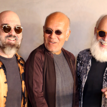 A imagem mostra os músicos Carlos Malta, Marcos Suzano e Alex Meirelles sorrindo em um clima descontraído. Todos usam óculos escuros e exibem visuais marcantes. Um deles tem barba cheia e veste uma camisa estampada em tons de preto e laranja, outro usa uma jaqueta marrom sobre uma camiseta cinza, e o terceiro tem uma longa barba branca e veste uma camisa estampada em preto e branco. A cena transmite cumplicidade e energia, refletindo a sintonia entre os artistas.