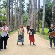 A imagem mostra as integrantes do grupo de forró Luz de Candeeiro em uma alameda cercada por palmeiras imperiais. Da esquerda para a direita, estão: Maria Souto tocando flauta, Marcela Coelho e Joana Araújo nas sanfonas, e Georgia Camara na percussão. As musicistas estão sorrindo e transmitindo alegria em meio ao cenário tropical, unindo natureza e cultura popular em uma atmosfera leve e vibrante.